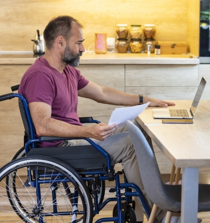 Handicapped mature guy in wheelchair working online from home, using laptop, looking for job opportunities on internet. Paraplegic mature man having business conference, making business project on web